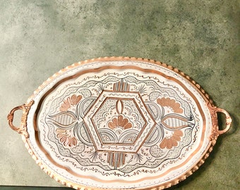 May include: An oval copper serving tray with a white background and intricate copper and silver floral designs. The tray has two handles on the sides.