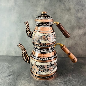 Può includere: Teiera in rame a due livelli con un design decorativo. La teiera presenta una finitura in rame con motivi floreali neri e bianchi. Ha un manico in legno e un beccuccio con un design nero e rame. Il coperchio è in rame con una parte superiore decorativa.
