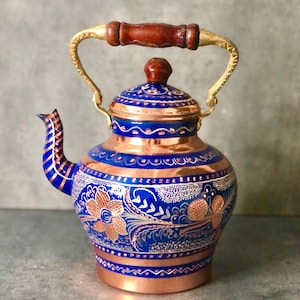 Può includere: Una teiera blu e rame con un manico dorato e intricati disegni floreali. La teiera ha un corpo rotondo e un coperchio con un pomo.