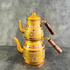 May include: A traditional Turkish tea set with two copper kettles, one stacked on top of the other. The kettles are decorated with intricate floral patterns in gold and have wooden handles.