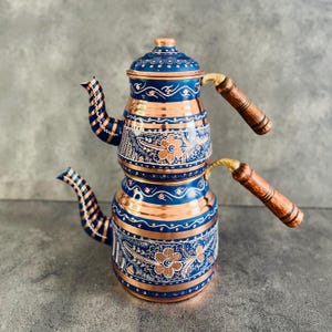 May include: A traditional Turkish tea kettle with a copper base and blue enamel. The kettle has a decorative floral design and two wooden handles.