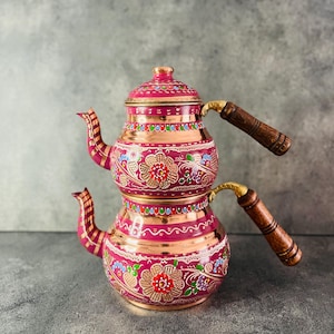 Può includere: Un set da tè turco tradizionale a due livelli. La teiera è in rame e decorata con motivi floreali dipinti a mano in tonalità di rosa, rosso, blu e verde. La teiera ha due manici in legno.