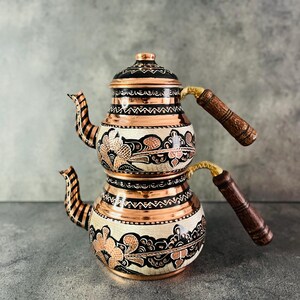Può includere: Una teiera in rame con un design floreale decorativo. La teiera presenta un motivo floreale in bianco e nero con accenti in rame. È dotata di un manico in legno e di un beccuccio con un motivo a strisce.