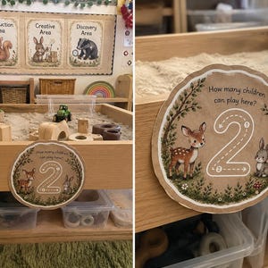 May include: A wooden sensory table filled with sand and small toys, featuring a circular sign with a deer, a rabbit, and the text "How many children can play here?" with the number 2, indicating the capacity for two children to play.