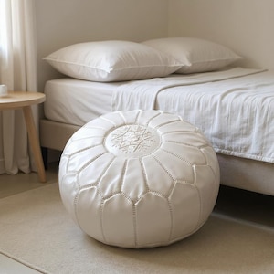 May include: A white, round Moroccan pouf with a geometric pattern and decorative stitching. The pouf is placed on a light-colored rug in front of a bed with white bedding. The pouf is a decorative and functional piece of furniture.