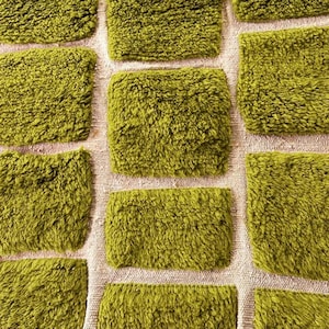 May include: Close-up of a textured, olive green fabric with a grid pattern. The squares are plush and fluffy, contrasting with the neutral background. The design suggests a rug or textile.