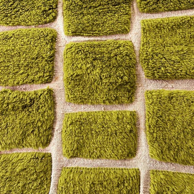 May include: Close-up of a textured, olive green fabric with a grid pattern. The squares are plush and fluffy, contrasting with the neutral background. The design suggests a rug or textile.