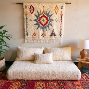 Puede incluir: Una cama baja de color crema con tres almohadas a juego y una pequeña almohada cuadrada. Una decoración mural decorativa con un diseño geométrico cuelga sobre la cama. Una alfombra estampada y plantas completan la decoración de la habitación.