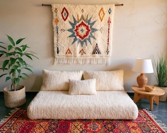 Op de afbeelding: Een crèmekleurig, laag bed met drie bijpassende kussens en een klein vierkant kussen. Boven het bed hangt een decoratieve wandhanger met een geometrisch ontwerp. Een patroonkleed en planten maken de kamerdecoratie compleet.
