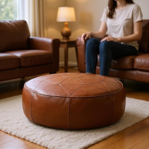 May include: A circular, tan leather ottoman with stitched geometric detailing. The ottoman is placed on a cream rug in a living room. A brown leather sofa and chair are in the background.