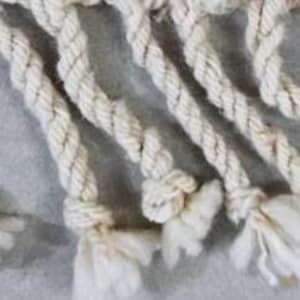 May include: Close-up of off-white macrame rope with intricate knots and fringed ends. The rope is tightly twisted, creating a textured surface. The image highlights the craftsmanship and detail of the handmade textile.