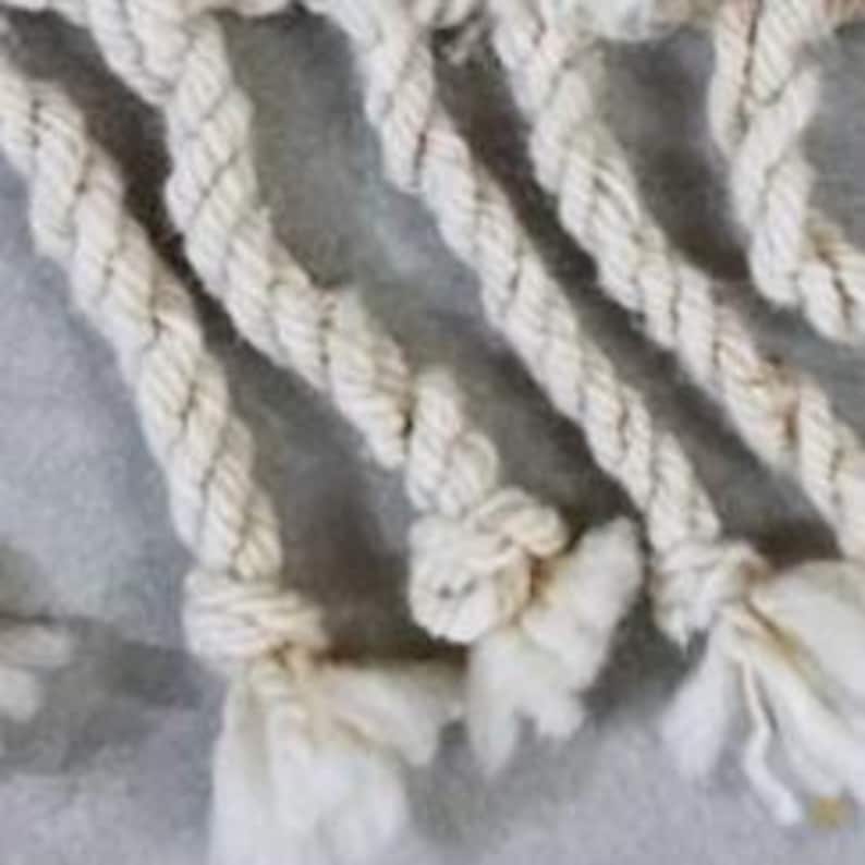 May include: Close-up of off-white macrame rope with intricate knots and fringed ends. The rope is tightly twisted, creating a textured surface. The image highlights the craftsmanship and detail of the handmade textile.