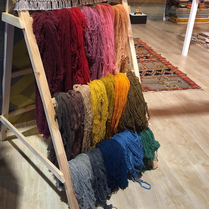 K&ouml;nnte beinhalten: Ein Holzregal zeigt B&uuml;ndel farbigen Garns in Rot-, Rosa-, Gelb-, Orange-, Blau- und Gr&uuml;nt&ouml;nen. Das Garn ist in vertikalen Reihen angeordnet und zeigt eine Vielzahl von Farbt&ouml;nen und Texturen. Das Regal ist aus hellem Holz gefertigt.