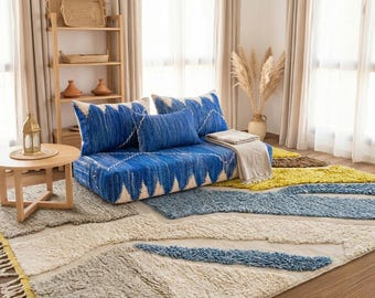 Op de afbeelding: Een gezellige woonkamer met een blauw en wit patroonbank en bijpassende kussens. Een kleine houten tafel heeft een lantaarn. De kamer is ingericht met tapijten in blauw, geel en beige, wat een warme en uitnodigende sfeer creëert.