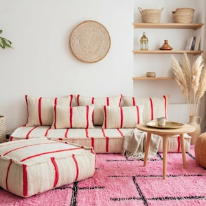 May include: A cozy living room setup featuring a cream-colored sofa and ottoman with red stripes, complemented by a pink rug. Decorative elements include a round wooden side table, a woven basket, and a potted plant.