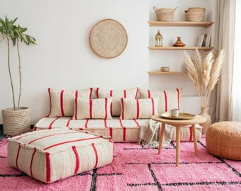 Op de afbeelding: Een gezellige woonkamer met een crèmekleurige bank en poef met rode strepen, op een roze tapijt. Een ronde houten tafel, een gevlochten mand en een potplant creëren een warme sfeer.