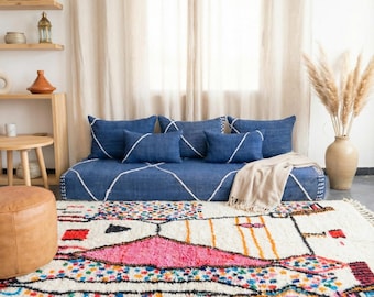 Op de afbeelding: Een woonkamerscène met een blauwe bank met witte geometrische lijnen en bijpassende kussens. Voor de bank ligt een kleurrijk tapijt met een geometrisch patroon. Een beige deken en een vaas met hoog gras maken de inrichting compleet.