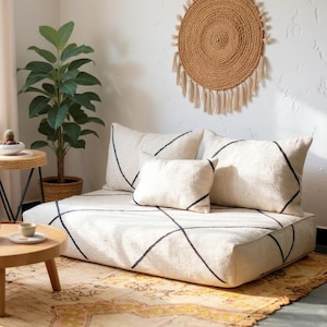 Op de afbeelding: Een woonkamer met een lage, vierkante bank met een crèmekleurige basis en zwarte geometrische lijnen. Crèmekleurige kussens met zwarte accenten en een klein rechthoekig kussen zijn op de bank geplaatst. Decoratieve planten en een ronde wanddecoratie.