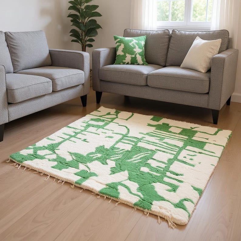 K&ouml;nnte beinhalten: Ein rechteckiger Teppich mit cremefarbenem Hintergrund und einem gr&uuml;nen, abstrakten Muster. Der Teppich liegt auf einem Holzboden vor einem grauen Sofa und einem Sessel. Ein gr&uuml;nes Kissen mit Muster liegt auf dem Sofa.