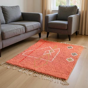 Pu&ograve; includere: Un tappeto rettangolare color corallo con un design geometrico bianco, rosso e oro. Il tappeto ha una superficie strutturata e frange bianche lungo i bordi. &Egrave; posizionato su un pavimento in legno chiaro.