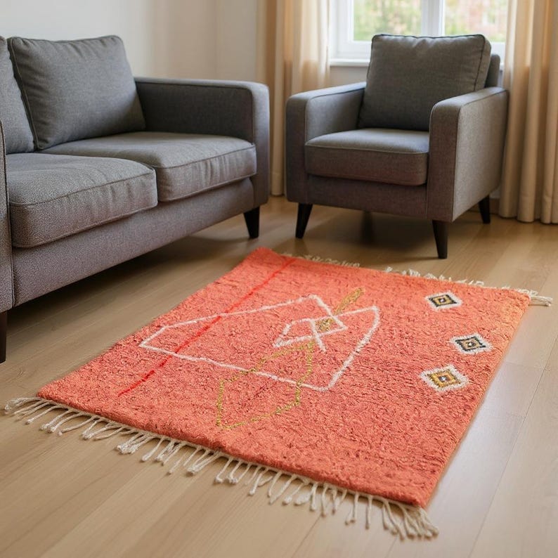 Pu&ograve; includere: Un tappeto rettangolare color corallo con un design geometrico bianco, rosso e oro. Il tappeto ha una superficie strutturata e frange bianche lungo i bordi. &Egrave; posizionato su un pavimento in legno chiaro.