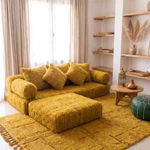 May include: A mustard yellow modular sofa with a matching rectangular ottoman and a textured shag rug. The sofa features two cylindrical side bolsters and three square back cushions, all covered in a shaggy, high-pile fabric material.