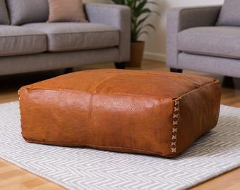 May include: A square, brown leather ottoman with visible stitching on the sides. The ottoman sits on a gray and white patterned rug, with a gray sofa and armchair in the background.