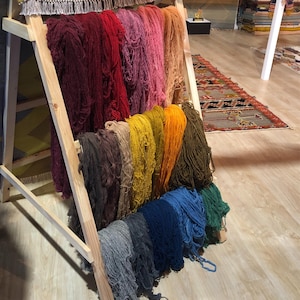 May include: A wooden display rack holds bundles of colorful yarn in shades of red, pink, yellow, orange, blue, and green. The yarn is arranged in vertical rows, showcasing a variety of hues and textures. The rack is made of light-colored wood.