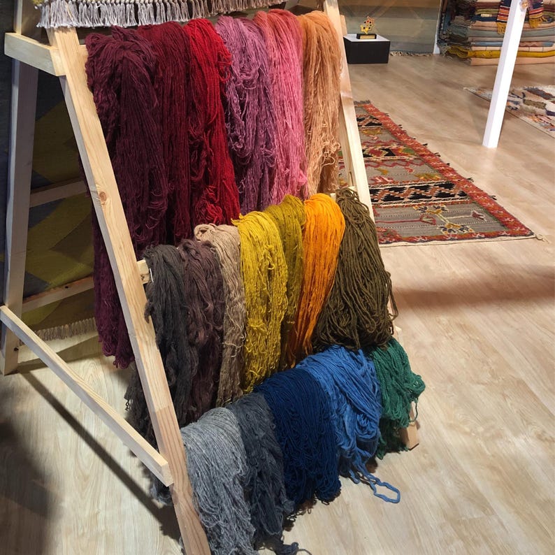 May include: A wooden display rack holds bundles of colorful yarn in shades of red, pink, yellow, orange, blue, and green. The yarn is arranged in vertical rows, showcasing a variety of hues and textures. The rack is made of light-colored wood.