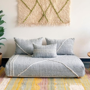 Puede incluir: Un cojín gris para el suelo con tres almohadas a juego, acentuado con costuras blancas. Los cojines están colocados sobre una alfombra colorida. Un tapiz de pared tejido y plantas en macetas completan la escena.