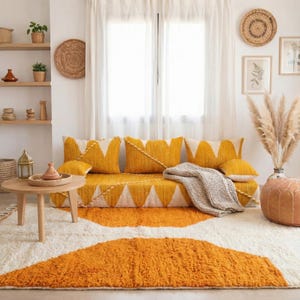 May include: A bright living room with a yellow sofa and cushions, featuring white triangle patterns. A cream rug with an orange design is in front of the sofa. The room is decorated with a round wooden table, woven wall art, and a tan leather pouffe.