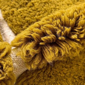 May include: Close-up of a mustard yellow, textured fabric. The fabric has a thick, shaggy pile, with visible strands and a soft, plush appearance. The material appears to be a rug or textile, with a neutral-colored backing.