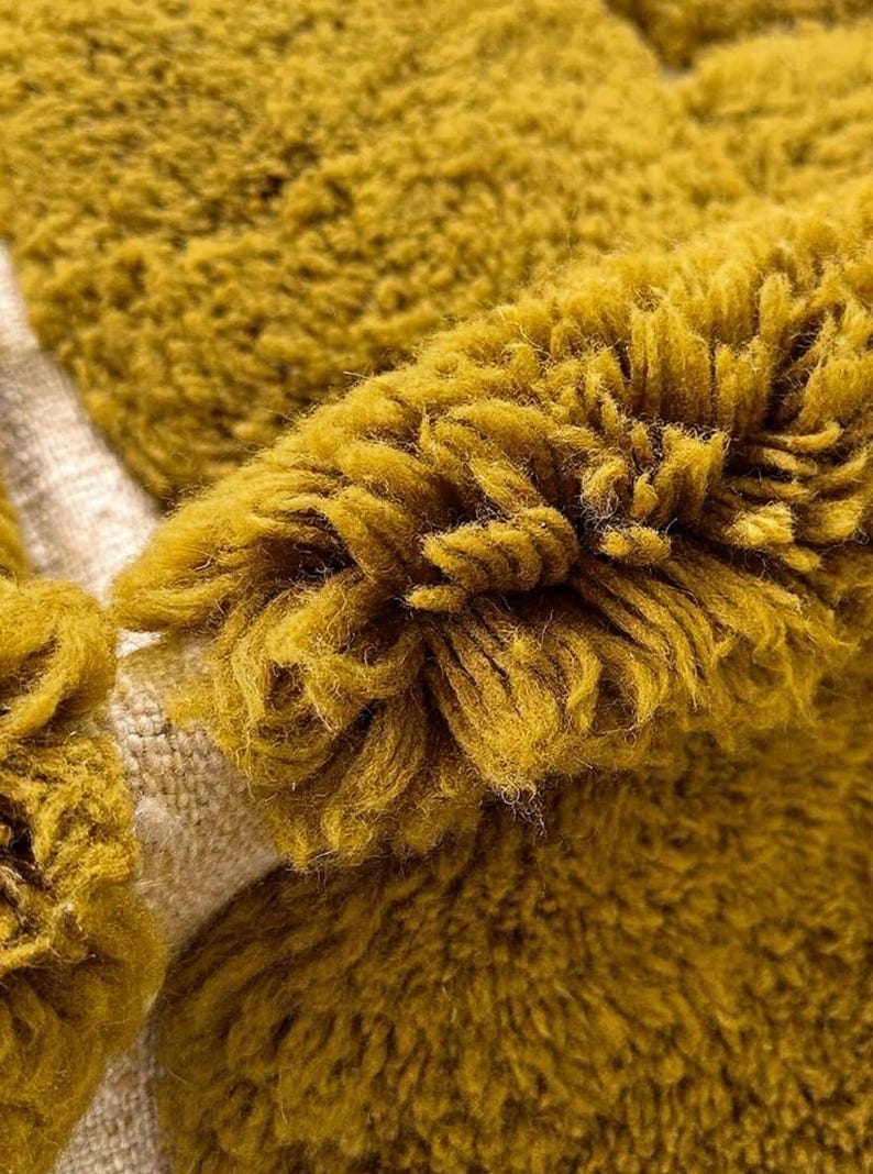 May include: Close-up of a mustard yellow, textured fabric. The fabric has a thick, shaggy pile, with visible strands and a soft, plush appearance. The material appears to be a rug or textile, with a neutral-colored backing.