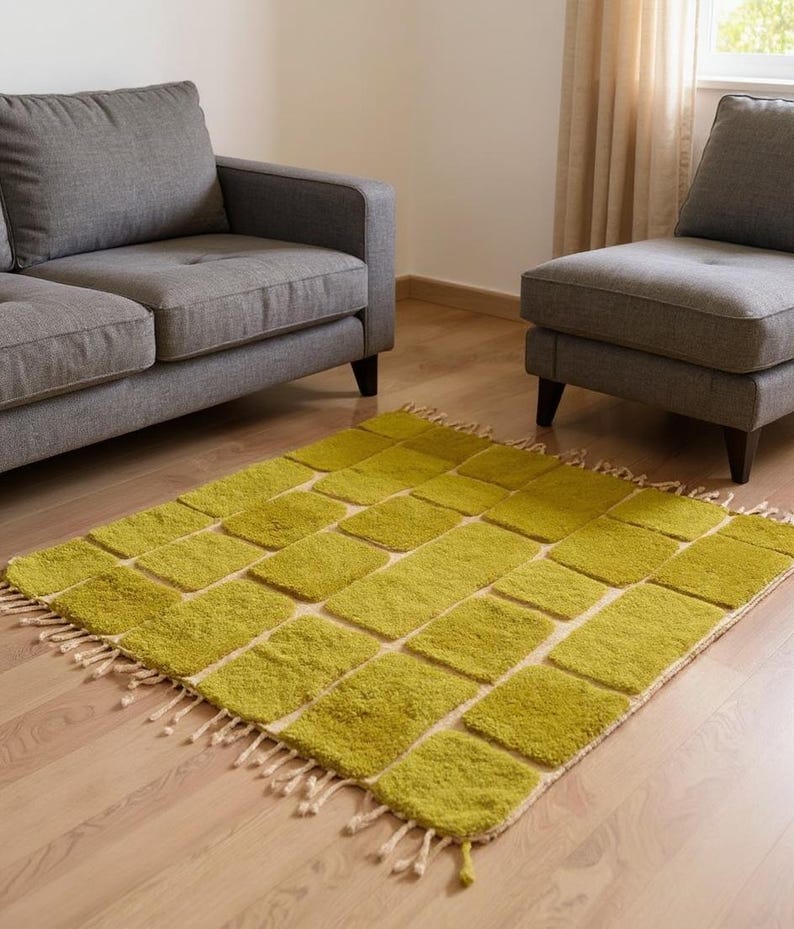 May include: A rectangular area rug with a geometric pattern of lime green squares on a cream background. The rug has a fringed edge and is placed on a hardwood floor. Two gray upholstered sofas are in the background.