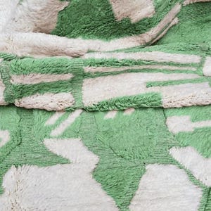 K&ouml;nnte beinhalten: Nahaufnahme eines weichen Teppichs mit einem geometrischen Muster in Gr&uuml;n und Off-White. Das Design zeigt rechteckige und abstrakte Formen, die einen optisch ansprechenden Textil schaffen. Die Textur des Teppichs ist weich.