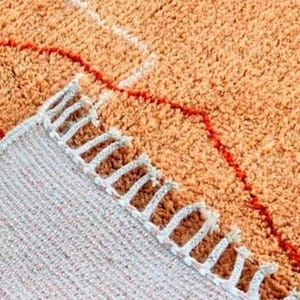 Pu&ograve; includere: Primo piano di un tappeto arancione strutturato con un design geometrico. Il tappeto presenta un motivo rosso e bianco e un bordo con frange. Le fibre del tappeto sono visibili, creando una texture morbida e invitante.