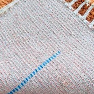 Pu&ograve; includere: Primo piano di una sezione di un tappeto, che mostra la trama strutturata e le variazioni di colore. Il tappeto ha una base grigio chiaro con macchie rosa e grigio pi&ugrave; scuro. Una linea blu attraversa il tappeto in diagonale.