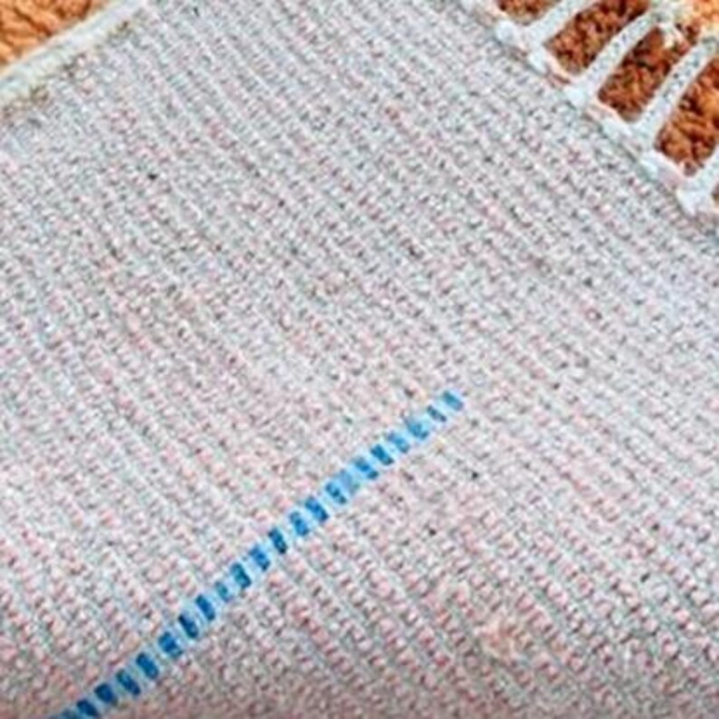 Pu&ograve; includere: Primo piano di una sezione di un tappeto, che mostra la trama strutturata e le variazioni di colore. Il tappeto ha una base grigio chiaro con macchie rosa e grigio pi&ugrave; scuro. Una linea blu attraversa il tappeto in diagonale.
