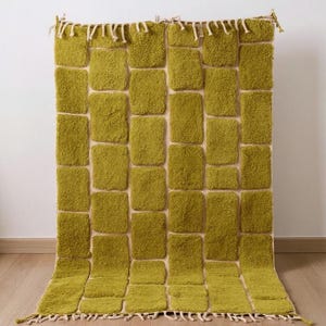 May include: A rectangular, olive green rug with a textured, brick-like pattern. The rug has a cream-colored background and a fringe along the bottom edge. The rug is displayed against a white wall and wooden floor.