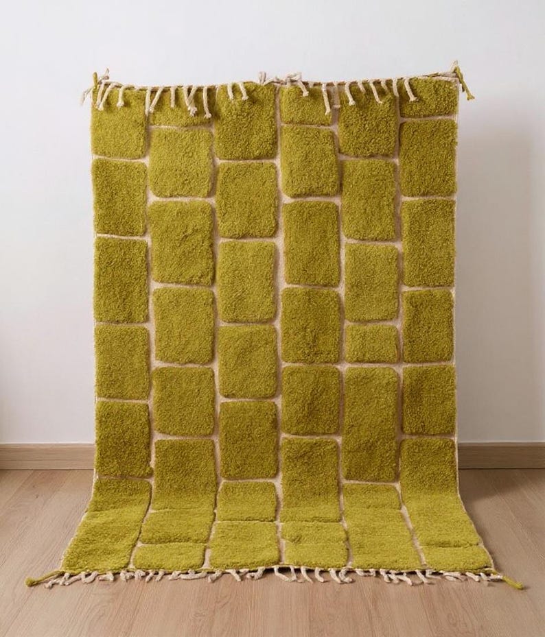 May include: A rectangular, olive green rug with a textured, brick-like pattern. The rug has a cream-colored background and a fringe along the bottom edge. The rug is displayed against a white wall and wooden floor.