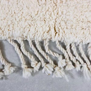 K&ouml;nnte beinhalten: Nahaufnahme eines cremefarbenen Teppichs mit einer strukturierten, zotteligen Oberfl&auml;che. Der Teppich hat einen Fransenbesatz aus gedrehtem, cremefarbenem Seil mit geknoteten Enden.