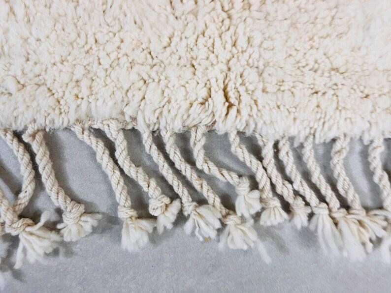 K&ouml;nnte beinhalten: Nahaufnahme eines cremefarbenen Teppichs mit einer strukturierten, zotteligen Oberfl&auml;che. Der Teppich hat einen Fransenbesatz aus gedrehtem, cremefarbenem Seil mit geknoteten Enden.