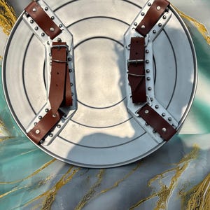May include: A silver-colored, round shield with brown leather straps and buckles. The shield has a central circular indentation and is reinforced with metal brackets and rivets. The straps are riveted to the shield's surface.