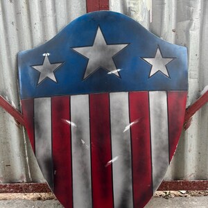 May include: A shield-shaped object with a blue upper section, displaying three silver stars. The lower part features red and white vertical stripes. The design is patriotic, similar to a superhero's emblem.