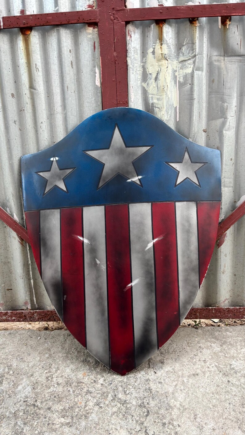 May include: A shield-shaped object with a blue upper section, displaying three silver stars. The lower part features red and white vertical stripes. The design is patriotic, similar to a superhero's emblem.