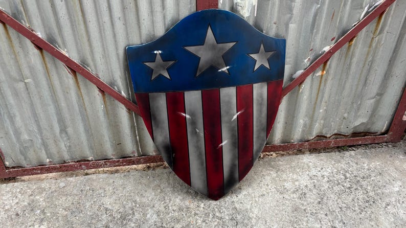 May include: A shield-shaped decorative item with a patriotic design. The top portion is blue with three silver stars, and the lower section features red and white stripes. The shield is displayed against a corrugated metal background.