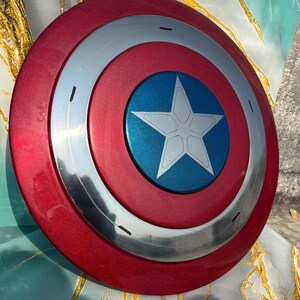 May include: A red, silver, and blue shield with a star emblem. The shield has a metallic finish and a circular design. The center features a blue circle with a white star. A classic superhero accessory.