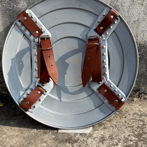 May include: A round, silver-colored shield with riveted metal supports and brown leather straps. The shield has a ridged design and is likely made of metal. The straps are secured with rivets.