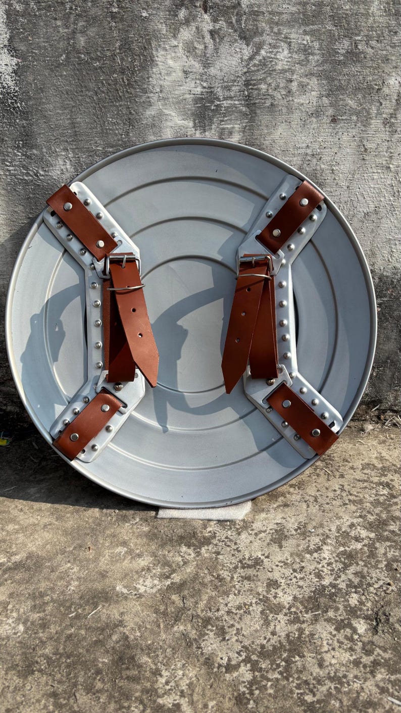 May include: A round, silver-colored shield with riveted metal supports and brown leather straps. The shield has a ridged design and is likely made of metal. The straps are secured with rivets.