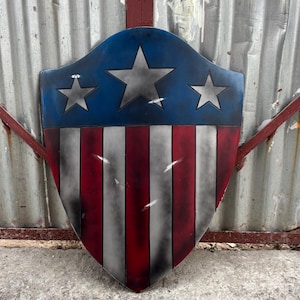 May include: A shield with a blue top section featuring three silver stars. The lower portion displays red and white stripes. The shield has a distressed, weathered appearance, suggesting a vintage or battle-worn aesthetic.